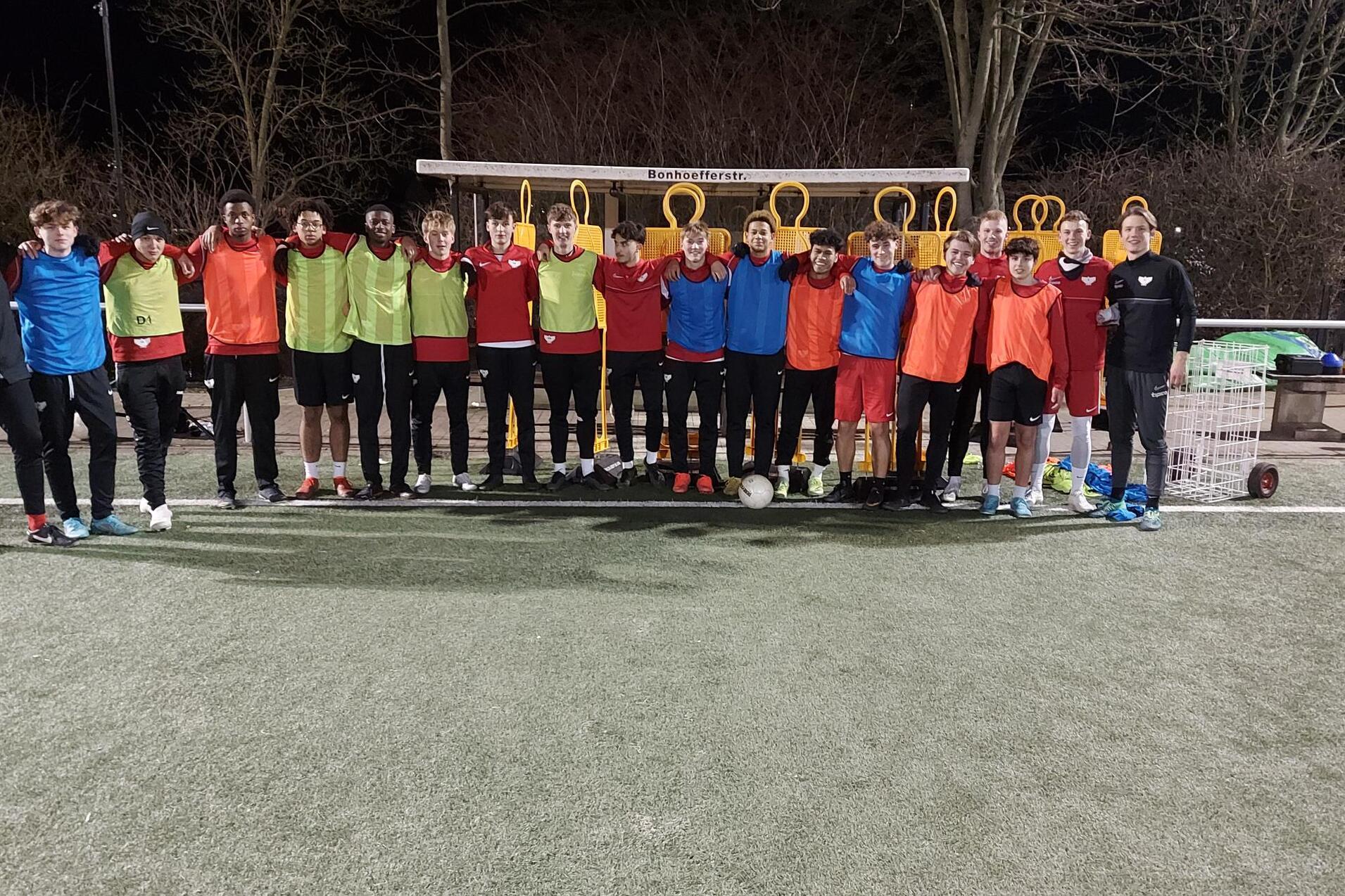 Gruppenzusammenstellung von 18 Fu&szlig;ballspielern in Trainingskleidung auf einem Kunstrasenplatz bei Nacht.