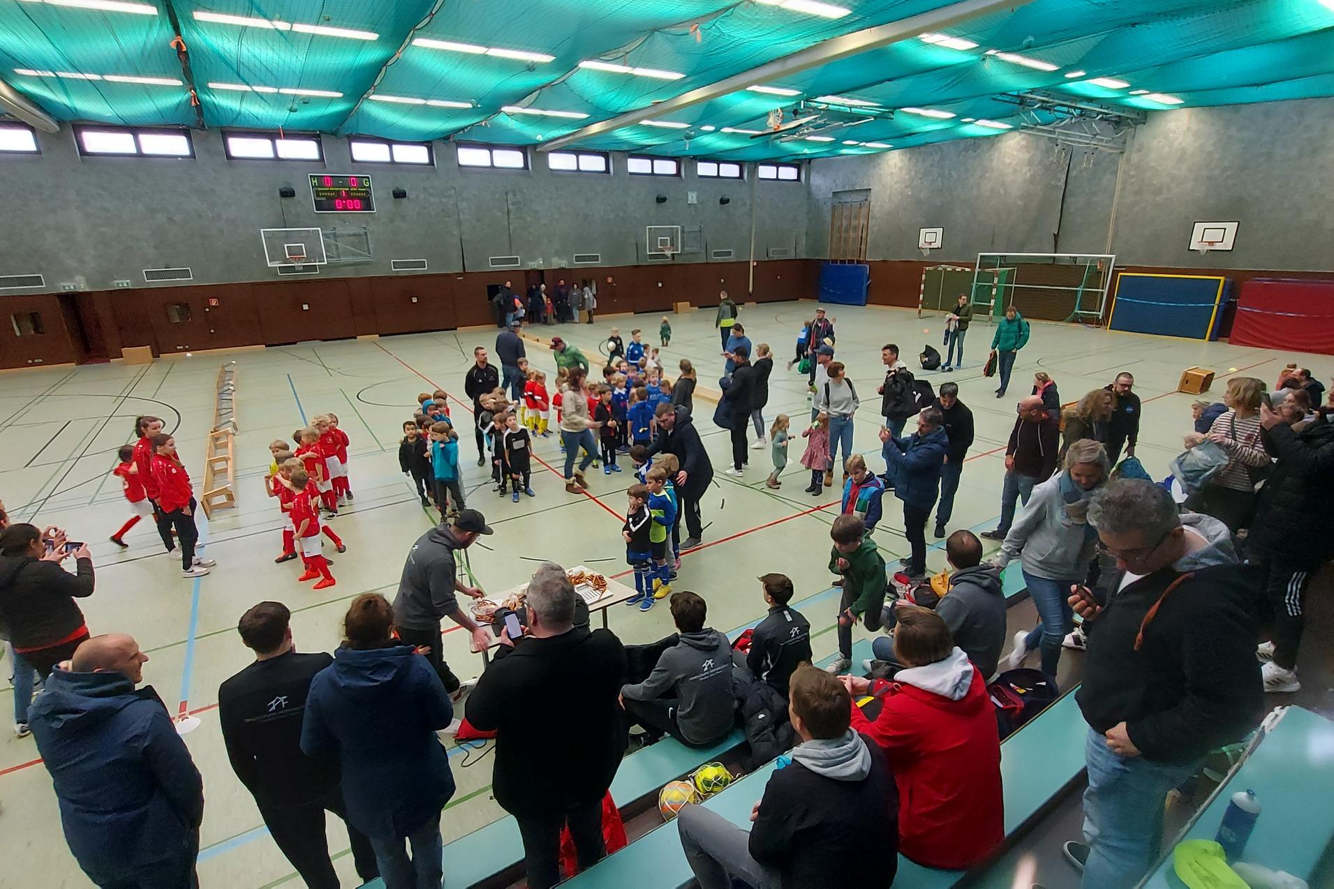 Ein lebhaftes Hallenfu&szlig;ballspiel mit Kindern, Zuschauern und Trainern in einer Sporthalle.