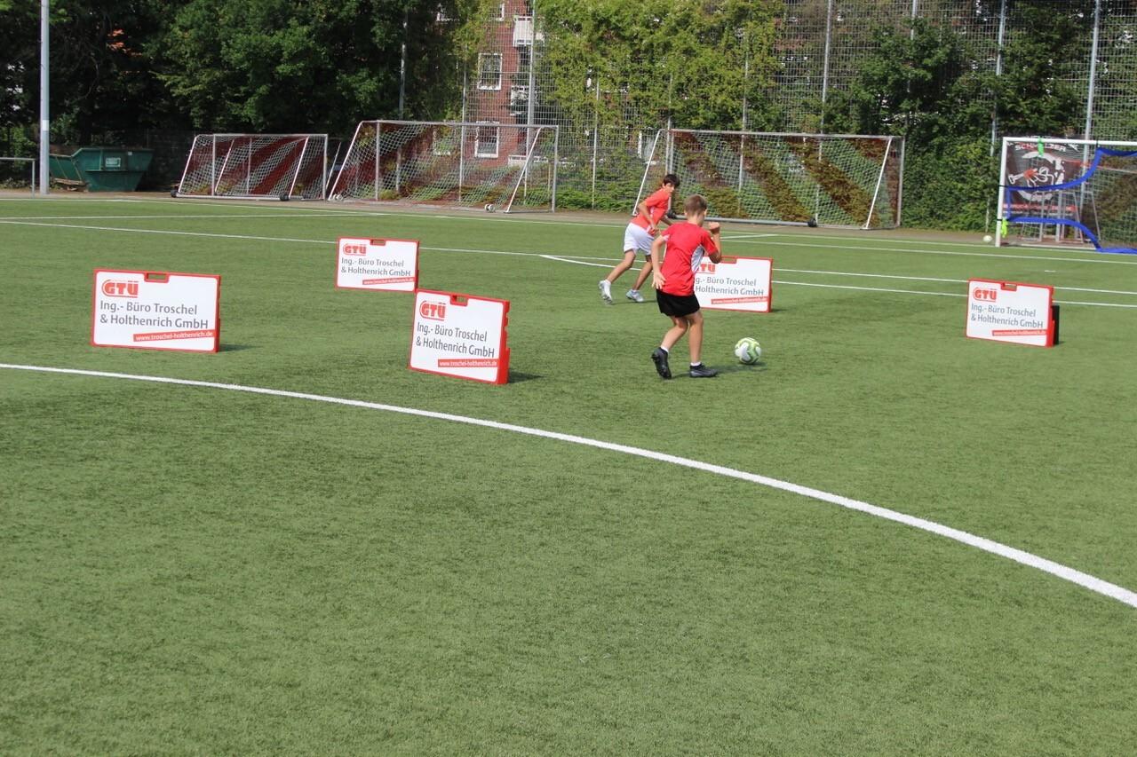Zwei Kinder spielen Fu&szlig;ball auf einem Kunstrasenplatz, umgeben von Werbeschildern.