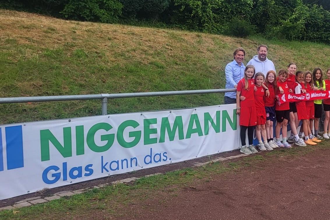 Gruppenshot von zwei Erwachsenen und neun M&auml;dchen in roten Trikots vor einem Sponsorenbanner am Fu&szlig;ballplatz.