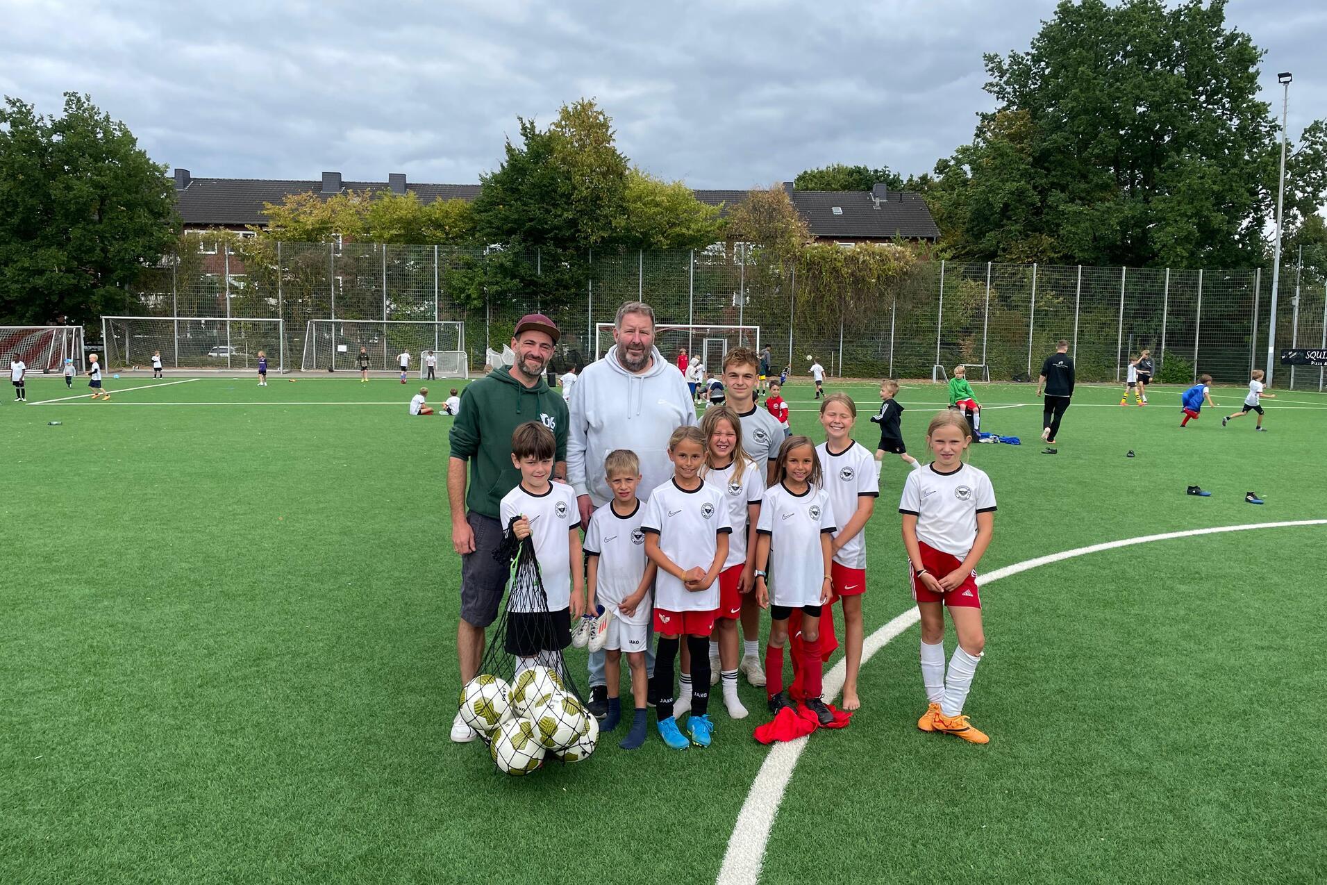 Gruppenfoto von zwei Trainern und neun Kindern in Sportkleidung auf einem Fu&szlig;ballfeld mit Ball.