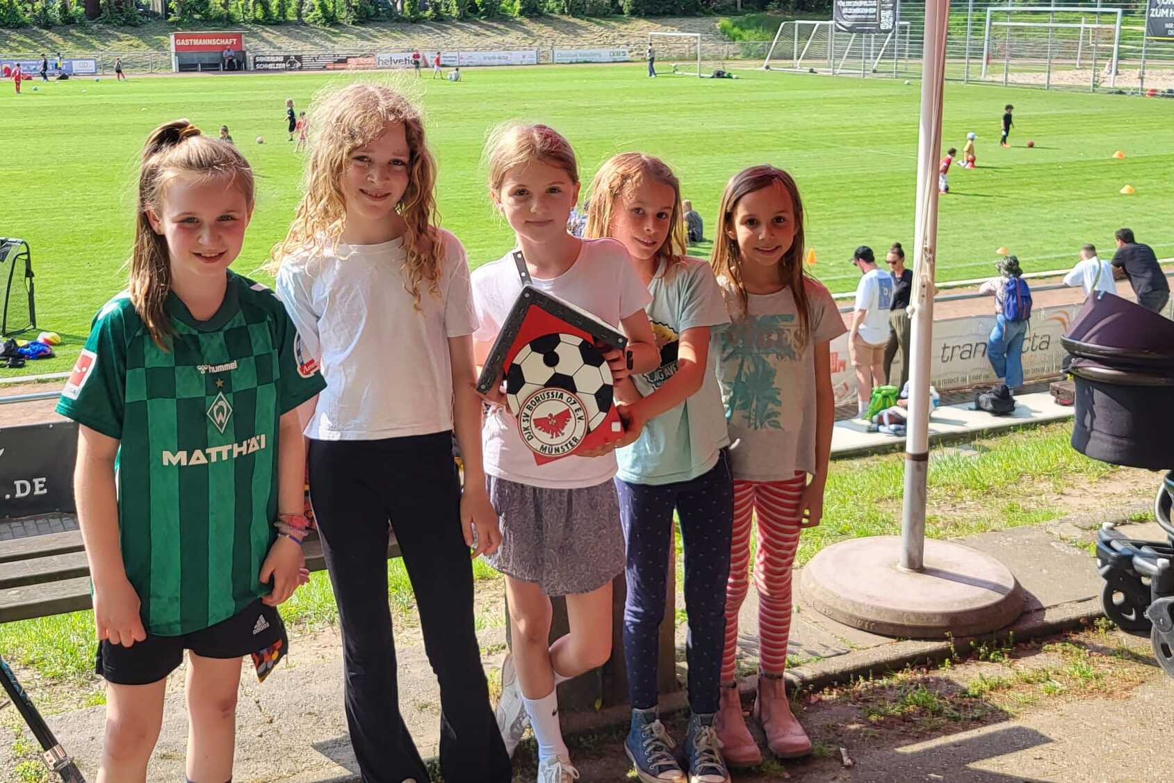 F&uuml;nf M&auml;dchen stehen l&auml;chelnd auf einem Sportplatz, halten ein Fu&szlig;ball-Troph&auml;e und tragen sportliche Kleidung.