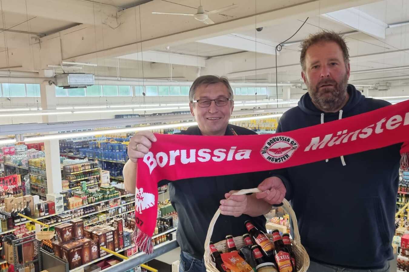 Zwei M&auml;nner halten einen Schal von Borussia M&uuml;nster und eine Getr&auml;nkekorb in einem Supermarkt. Vielen Regalen im Hintergrund.