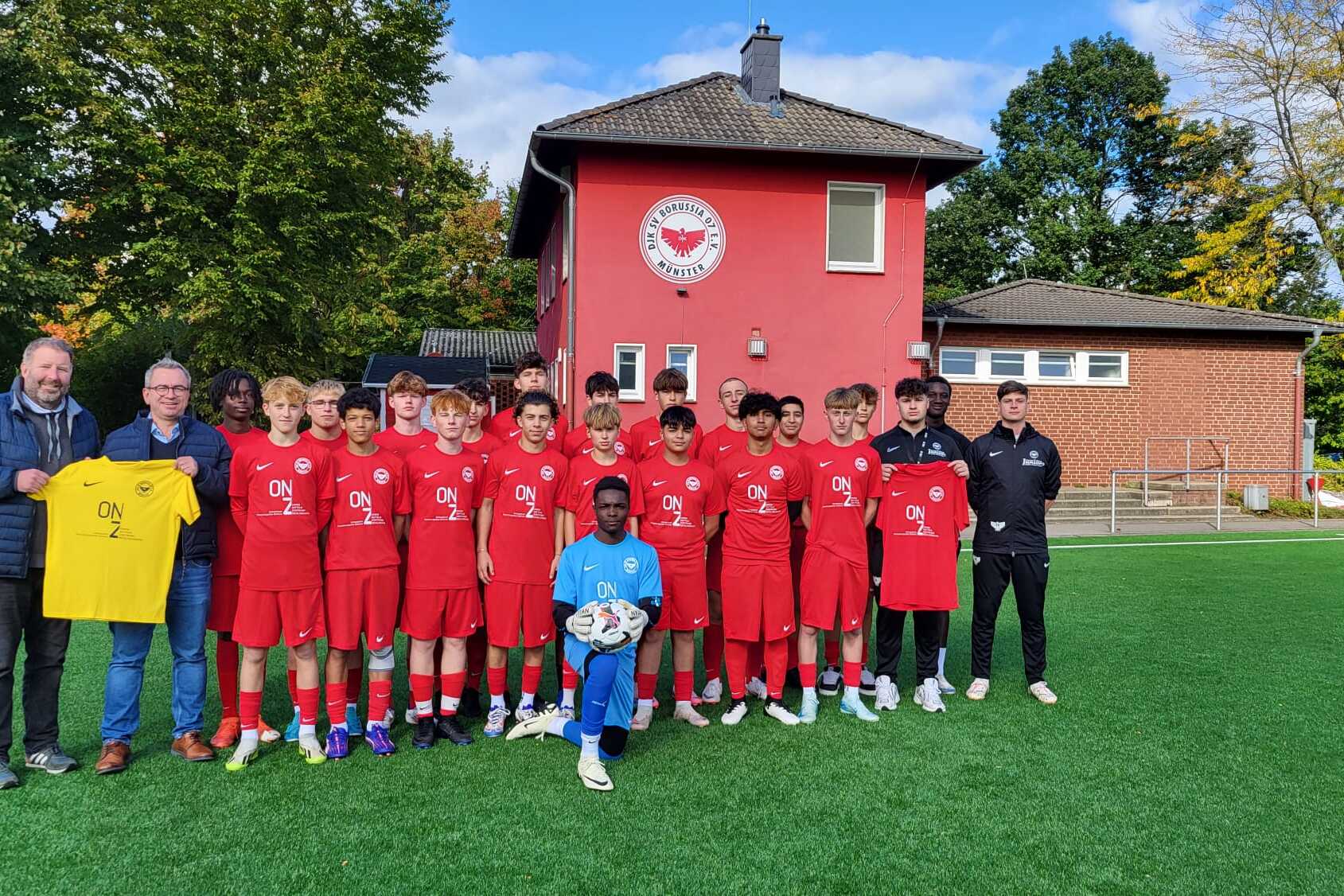 Jugendfu&szlig;ballmannschaft in roten Trikots steht vor einem Vereinsgeb&auml;ude, Trainer und Betreuer sind ebenfalls dabei.