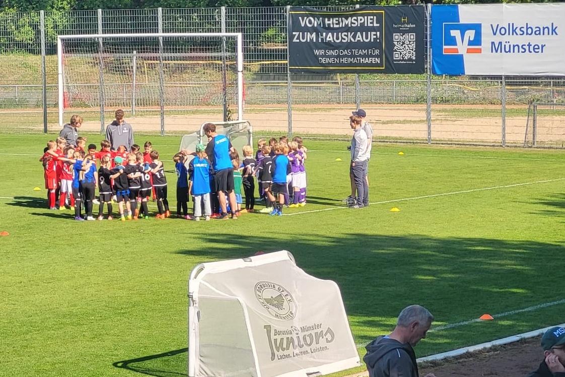 Gruppierung von Kindern in Sportkleidung auf einem Fu&szlig;ballplatz, umrahmt von Trainern und einer Werbebehinderung.
