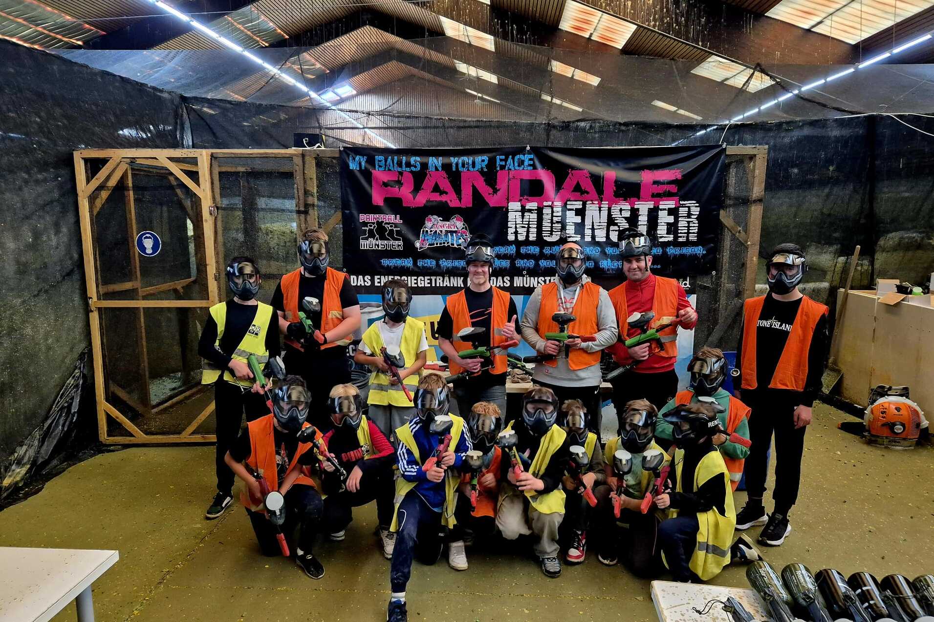 Gruppenselfie von Teilnehmern in Schutzkleidung mit Paintball-Ausr&uuml;stung vor einem Banner in einer Indoor-Spielarena.