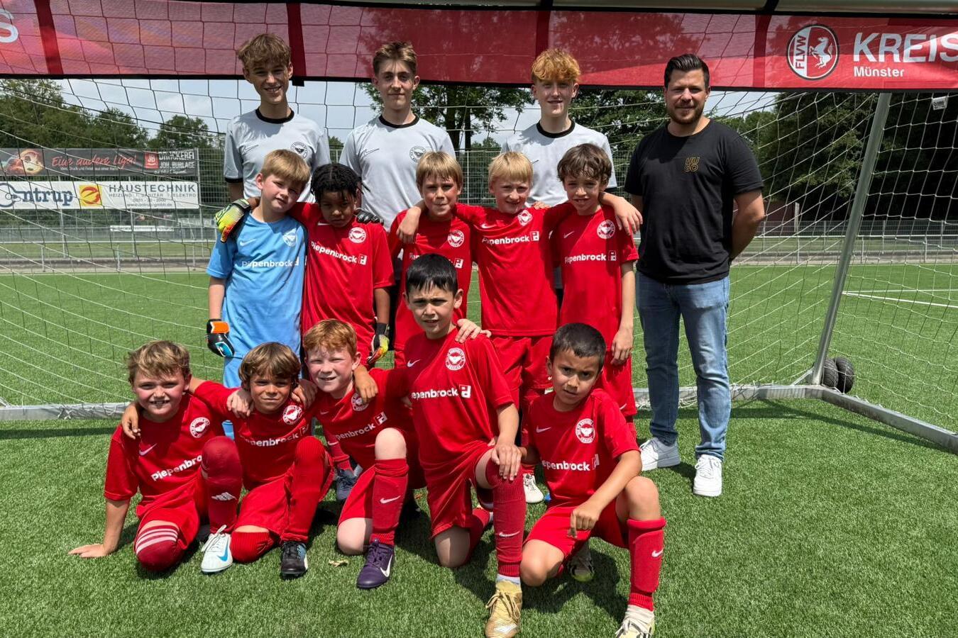 Gruppe von 13 jungen Fu&szlig;ballspielern in roten Trikots und zwei Trainern vor einem Tor auf einem Kunstrasenplatz.