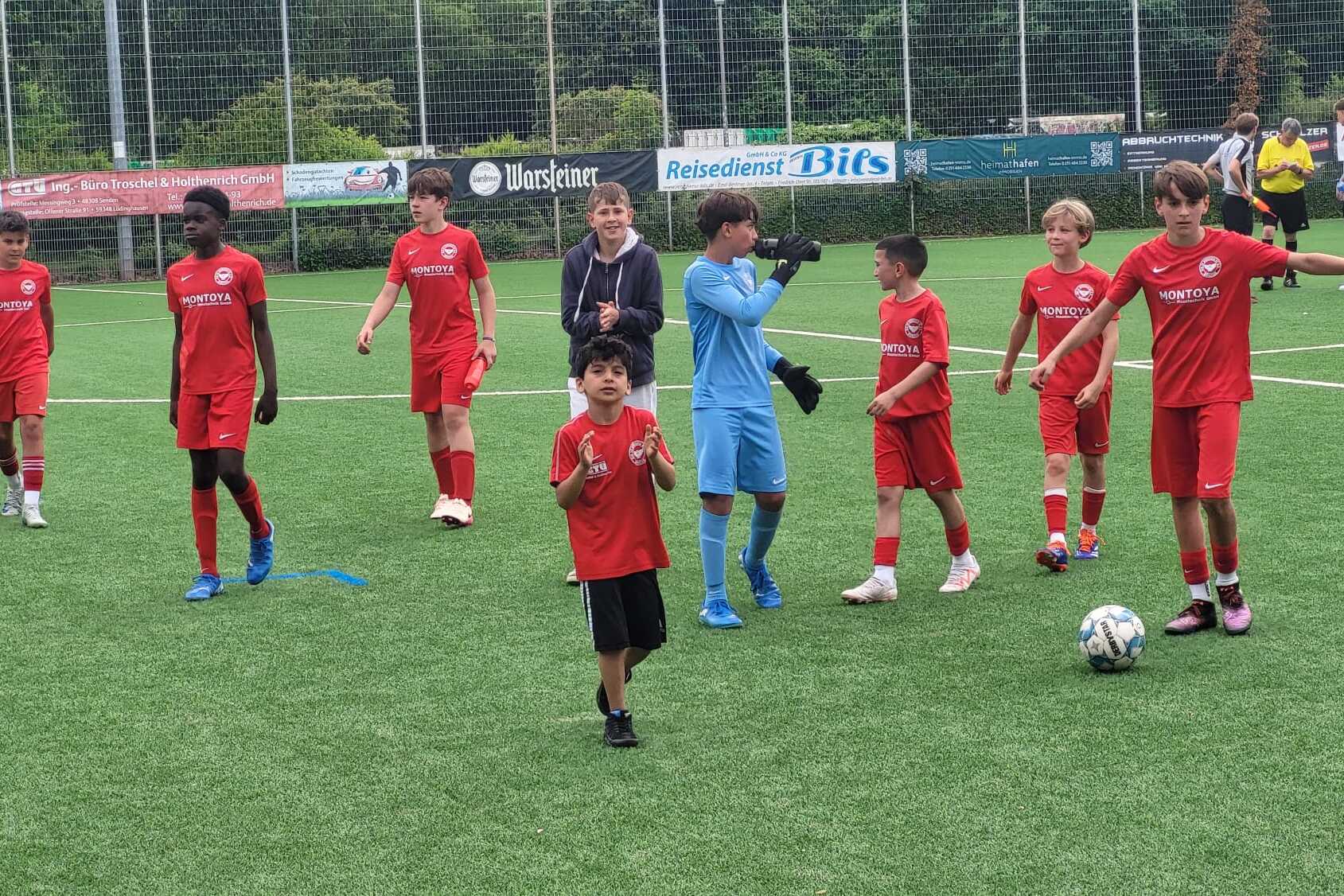 Gruppe von Kindern in roten Fu&szlig;balltrikots auf einem Kunstrasenplatz, einige stehen, andere laufen, ein Torwart in blauer Ausr&uuml;stung.