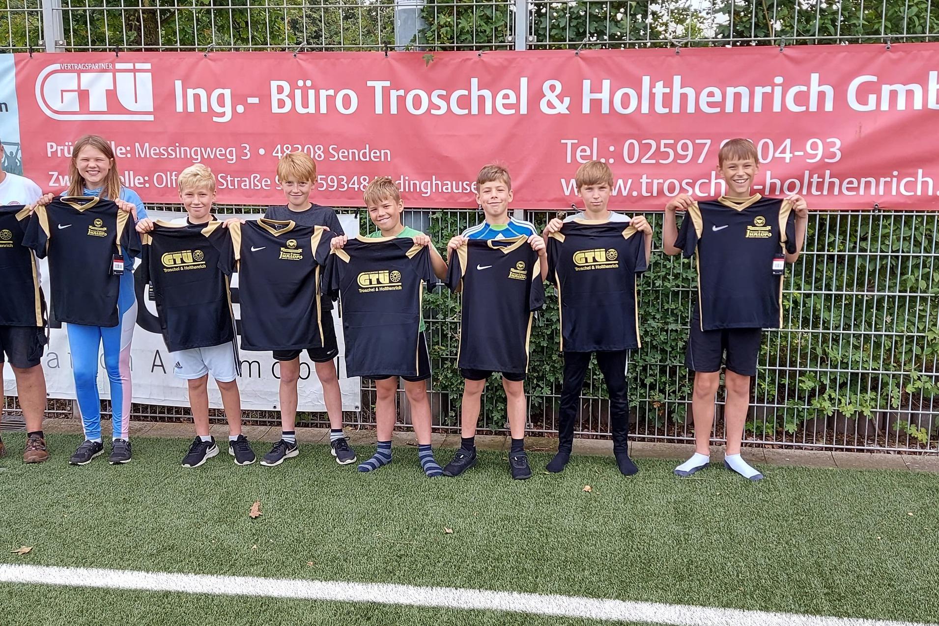 Gruppe von sieben Kindern in Sportbekleidung und einem Erwachsenen stehen vor einem Werbebanner auf einem Fußballfeld.