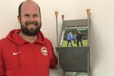 Mann in roter Sportjacke l&auml;chelt neben einem Regal mit Fu&szlig;balltrainingsmagazinen und -materialien.