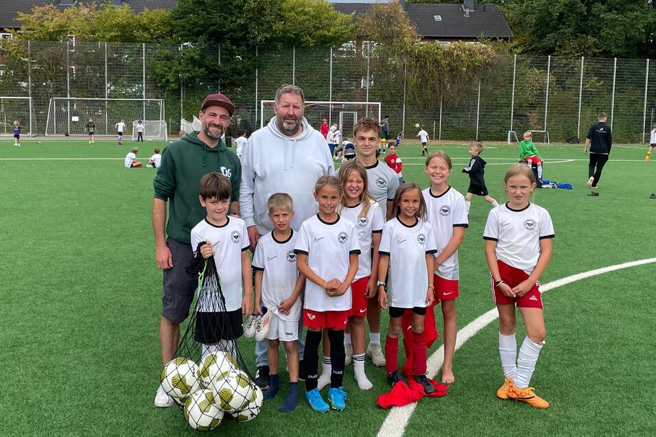 Gruppenfoto von zwei Trainern und neun Kindern in Sportkleidung auf einem Fu&szlig;ballfeld mit Ball.