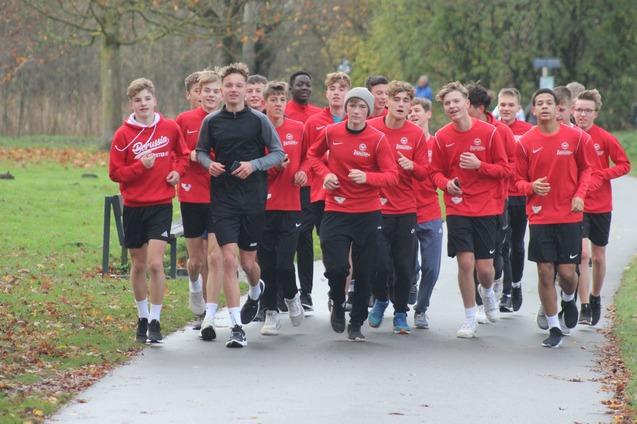 Gruppe von Jugendlichen in roten Trainingsshirts joggt auf einem Weg durch einen Park im Herbst.