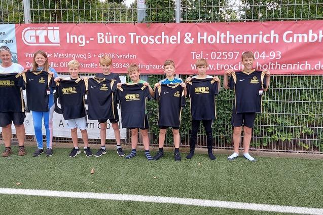Gruppe von sieben Kindern in Sportbekleidung und einem Erwachsenen stehen vor einem Werbebanner auf einem Fu&szlig;ballfeld.