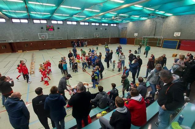 Ein lebhaftes Hallenfu&szlig;ballspiel mit Kindern, Zuschauern und Trainern in einer Sporthalle.