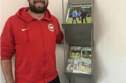 Mann in roter Sportjacke l&auml;chelt neben einem Regal mit Fu&szlig;balltrainingsmagazinen und -materialien.