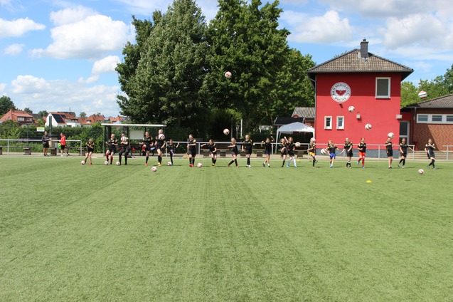 Gruppe von Fu&szlig;ballspielern in Training, die B&auml;lle auf einem Kunstrasenplatz passen, im Hintergrund ein rotes Vereinsgeb&auml;ude.