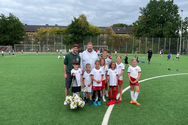 Gruppenfoto von zwei Trainern und neun Kindern in Sportkleidung auf einem Fu&szlig;ballfeld mit Ball.