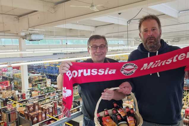 Zwei M&auml;nner halten einen Schal von Borussia M&uuml;nster und eine Getr&auml;nkekorb in einem Supermarkt. Vielen Regalen im Hintergrund.