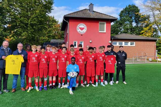 Jugendfu&szlig;ballmannschaft in roten Trikots steht vor einem Vereinsgeb&auml;ude, Trainer und Betreuer sind ebenfalls dabei.