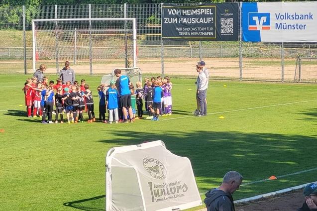 Gruppierung von Kindern in Sportkleidung auf einem Fu&szlig;ballplatz, umrahmt von Trainern und einer Werbebehinderung.