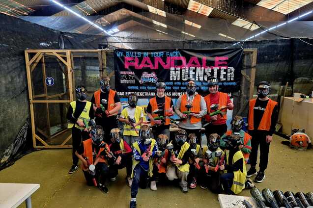 Gruppenselfie von Teilnehmern in Schutzkleidung mit Paintball-Ausr&uuml;stung vor einem Banner in einer Indoor-Spielarena.