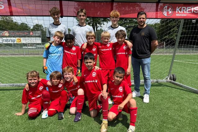Gruppe von 13 jungen Fu&szlig;ballspielern in roten Trikots und zwei Trainern vor einem Tor auf einem Kunstrasenplatz.