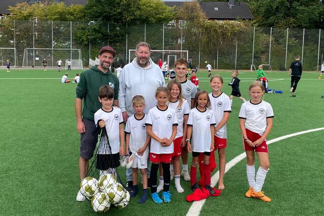 Gruppenfoto von zwei Trainern und neun Kindern in Sportkleidung auf einem Fu&szlig;ballfeld mit Ball.