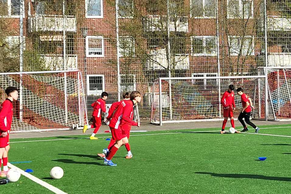 Gruppe von jungen Fu&szlig;ballspielern in roten Trikots trainiert auf einem Kunstrasenplatz mit zwei Toren im Hintergrund.