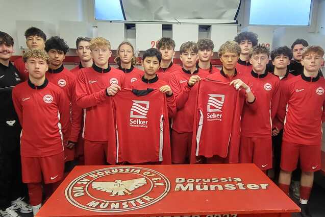 Gruppe von Fu&szlig;ballspielern in roten Trikots steht in einer Umkleidekabine, posierend mit neuen Trikots vor einem Tisch.