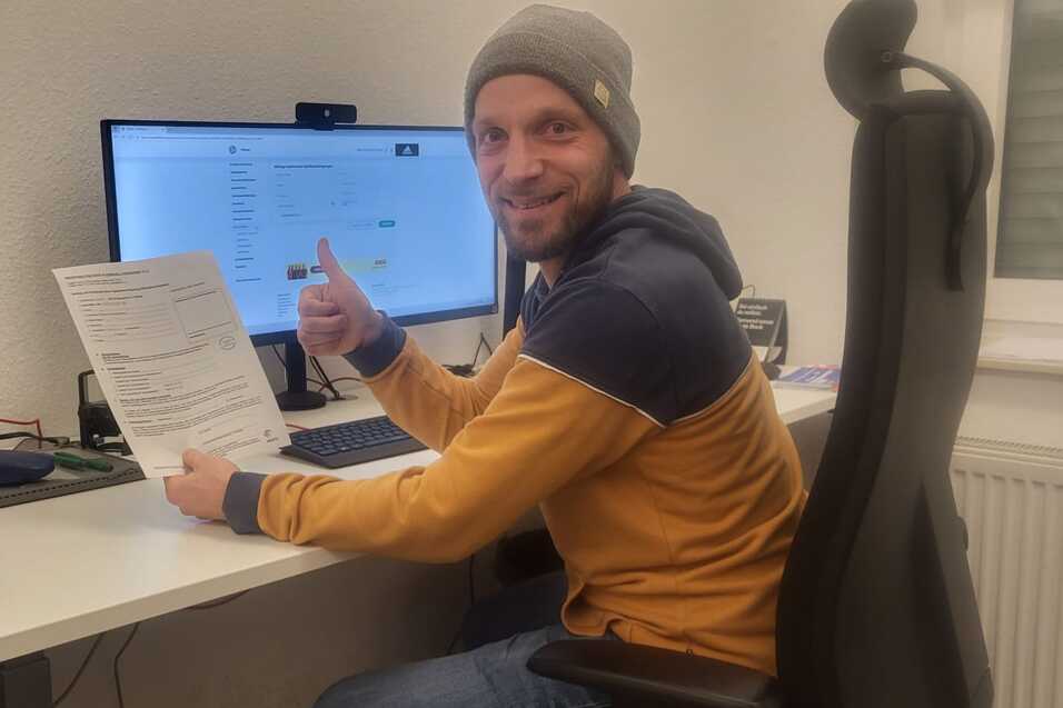 Mann in einem B&uuml;ro zeigt Daumen hoch und h&auml;lt ein Dokument in der Hand, sitzt vor einem Computer mit Monitor.