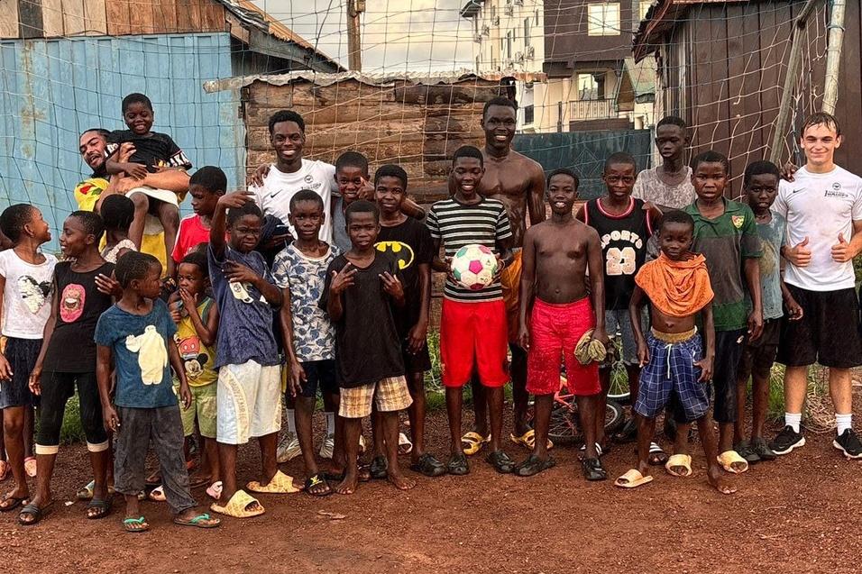 Gruppe von Kindern und Jugendlichen vor einem Fu&szlig;balltor, l&auml;chelnd und mit einem Ball, in einer l&auml;ndlichen Umgebung.