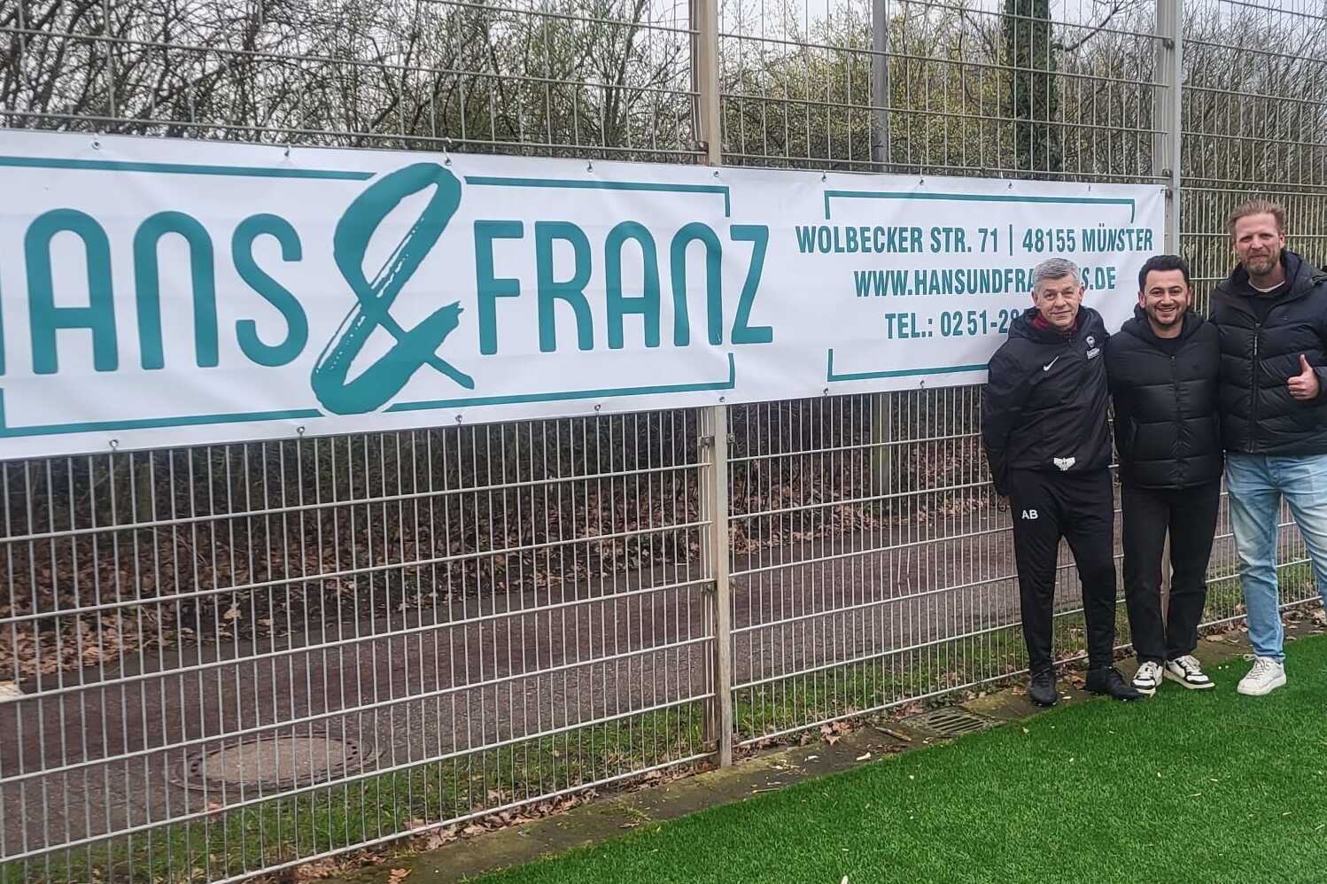 Drei Personen stehen vor einem Zaun mit einem Banner von "HANS & FRANZ" auf einem Fu&szlig;ballplatz.