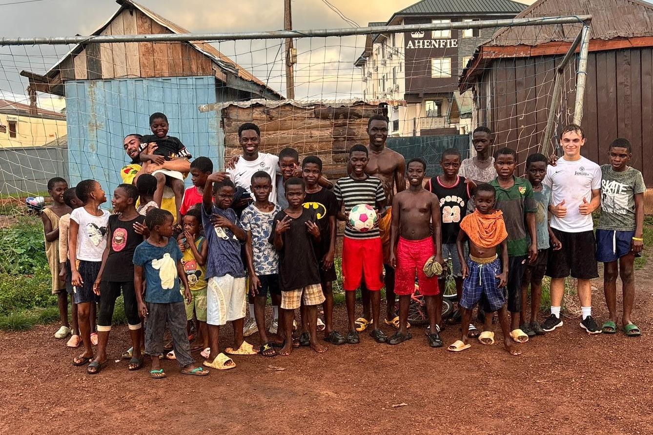 Gruppe von Kindern und Jugendlichen vor einem Fu&szlig;balltor, l&auml;chelnd und mit einem Ball, in einer l&auml;ndlichen Umgebung.