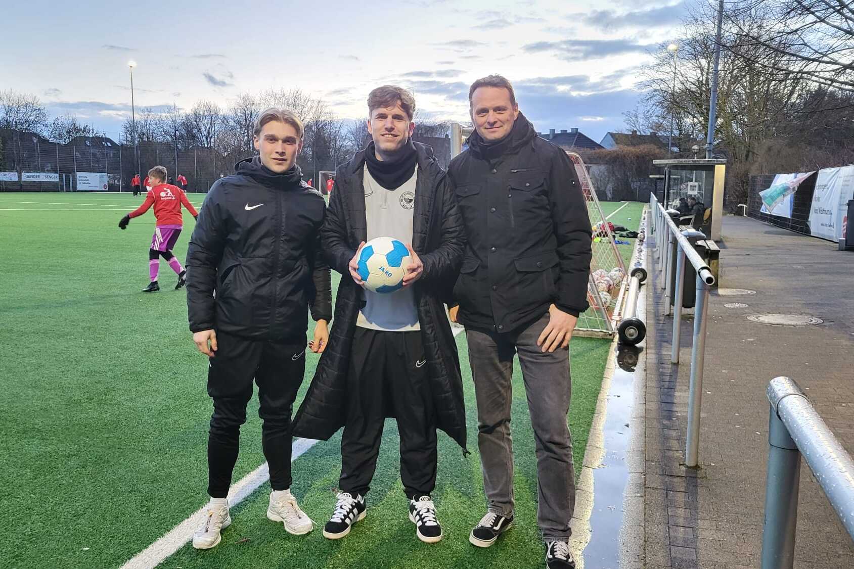 Drei Personen stehen auf einem Fu&szlig;ballplatz, eine h&auml;lt einen Ball, w&auml;hrend Spieler im Hintergrund aktiv sind.
