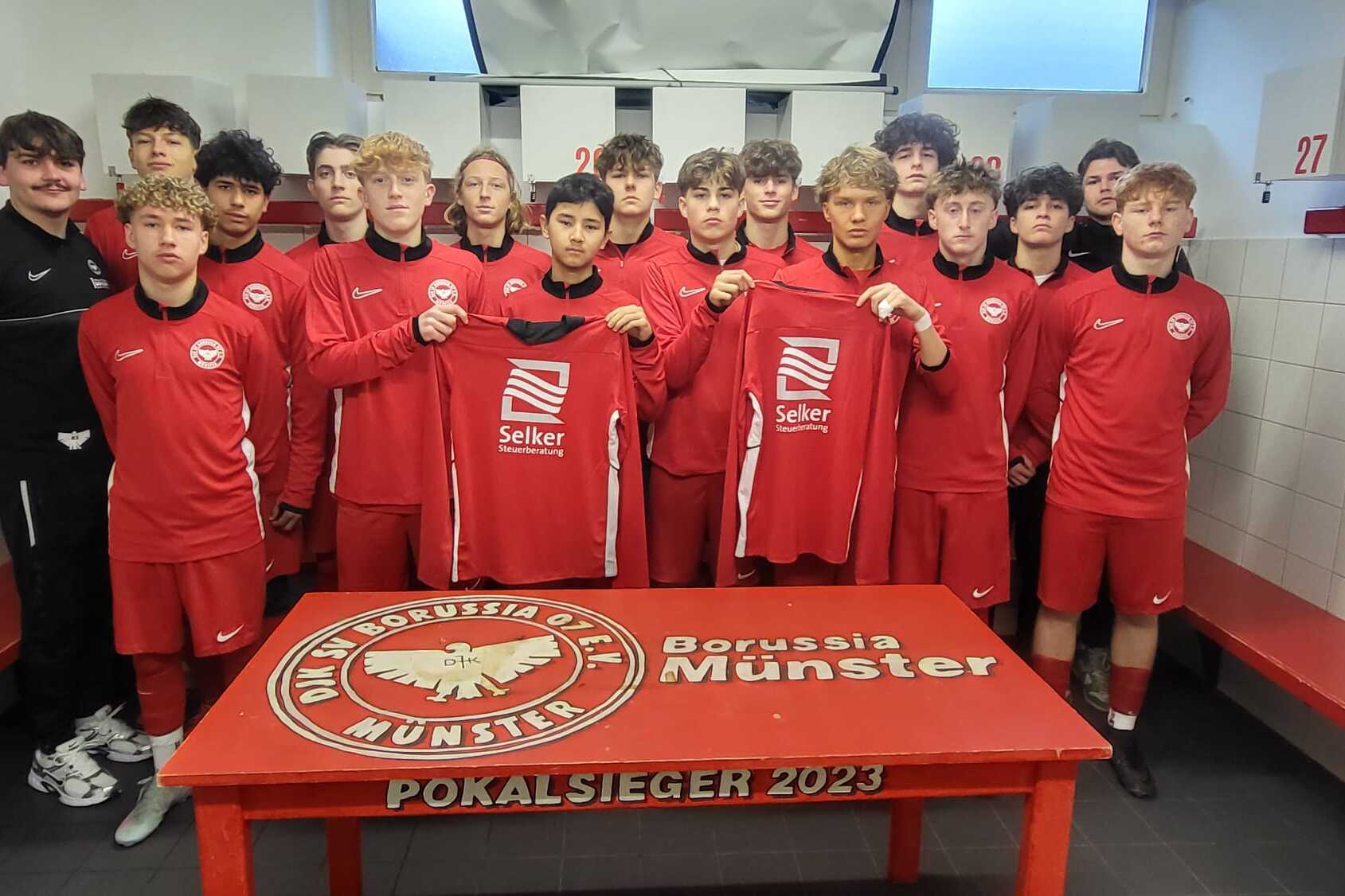 Gruppe von Fu&szlig;ballspielern in roten Trikots steht in einer Umkleidekabine, posierend mit neuen Trikots vor einem Tisch.