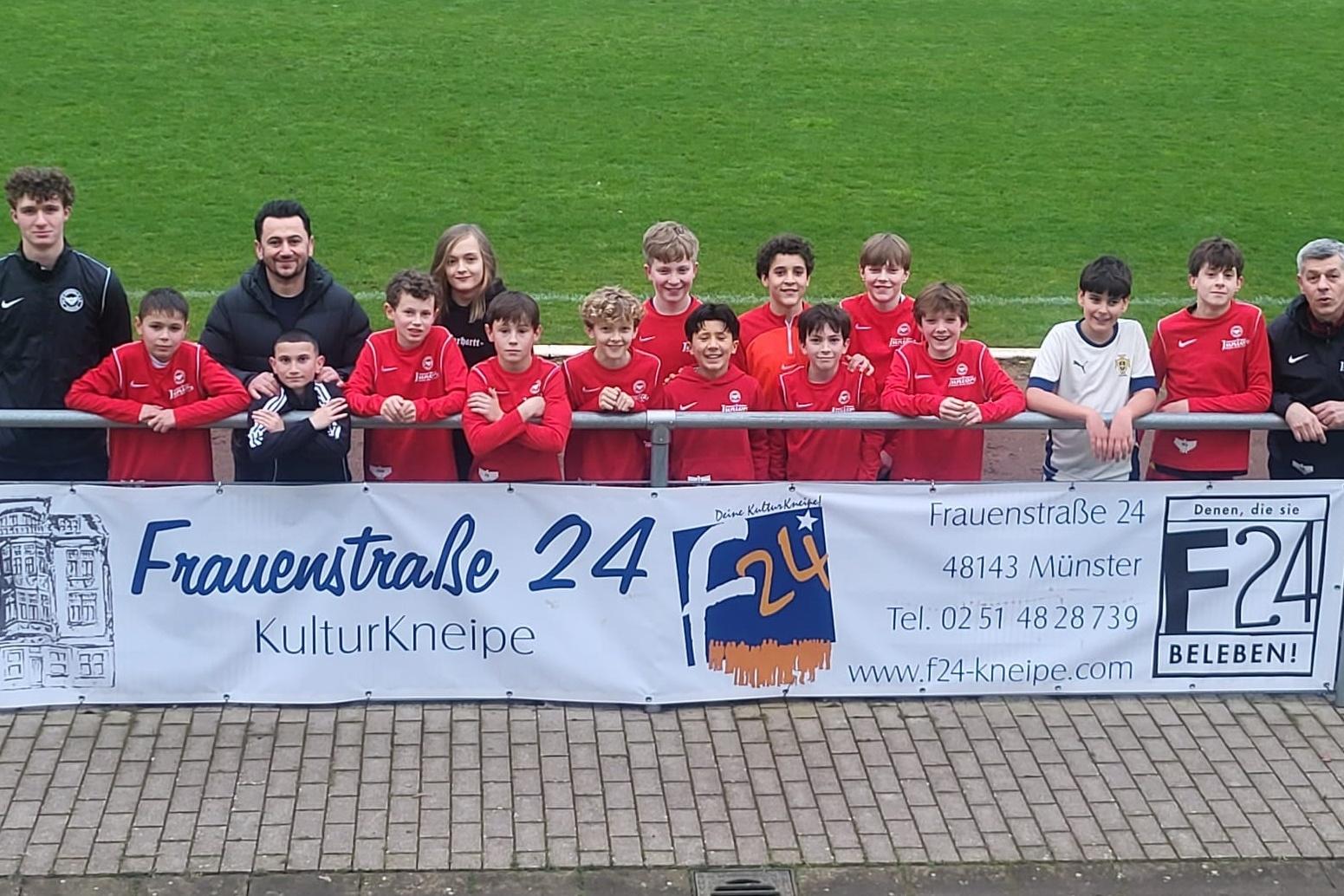 Gruppennfoto von Kindern und Trainern in roten Trikots vor einem Banner der Kulturkneipe Frauenstra&szlig;e 24.
