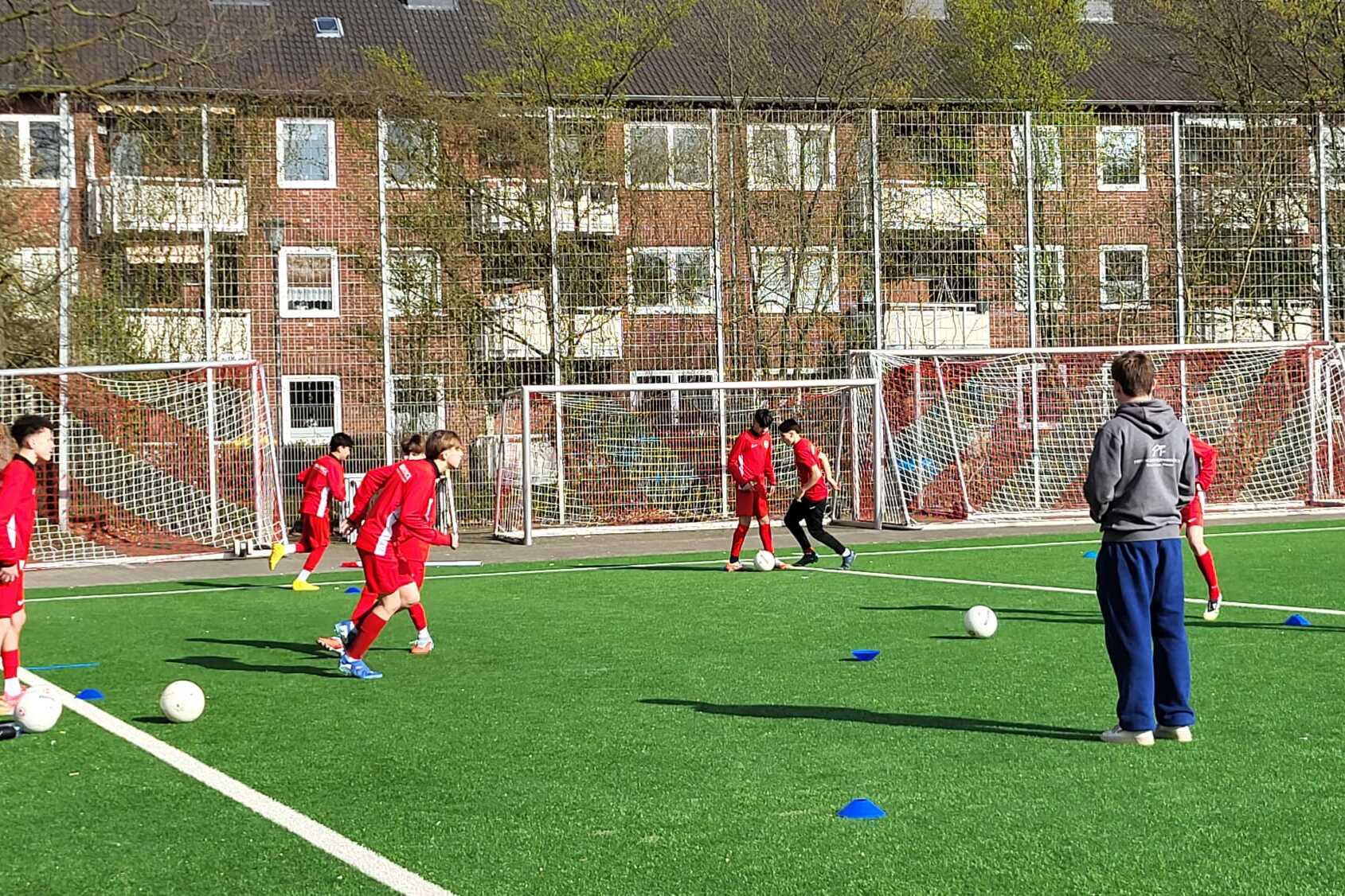 Gruppe von jungen Fu&szlig;ballspielern in roten Trikots trainiert auf einem Kunstrasenplatz mit zwei Toren im Hintergrund.