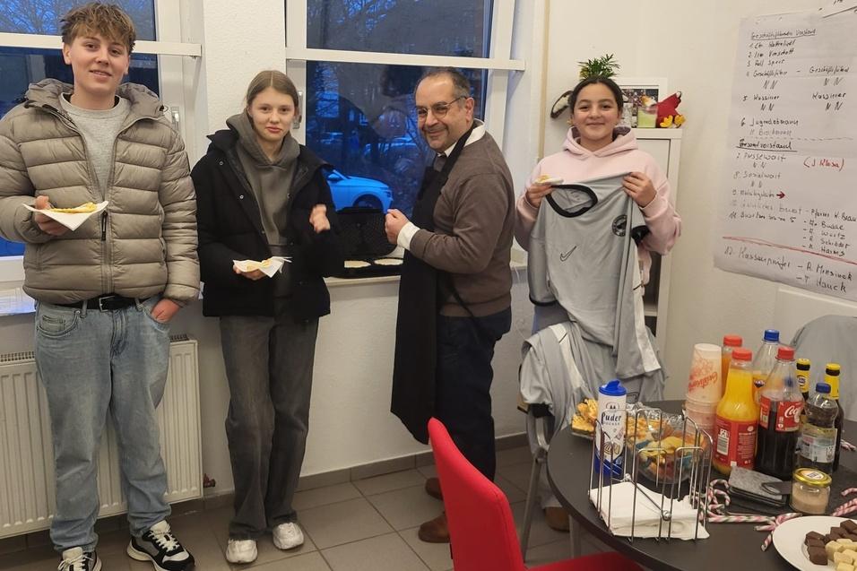 Vier Personen in einem Raum halten Essen und T-Shirts, w&auml;hrend einer eine Bestellung &uuml;bergibt. Hinter ihnen steht ein Tisch mit Snacks.