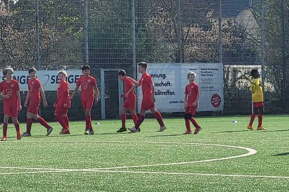 Sechs Fu&szlig;ballspieler in roten Trikots laufen auf einem Kunstrasenplatz, w&auml;hrend ein Spieler in blauem Trikot wartet.