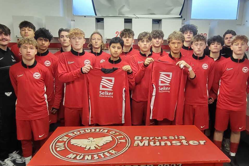 Gruppe von Fu&szlig;ballspielern in roten Trikots steht in einer Umkleidekabine, posierend mit neuen Trikots vor einem Tisch.