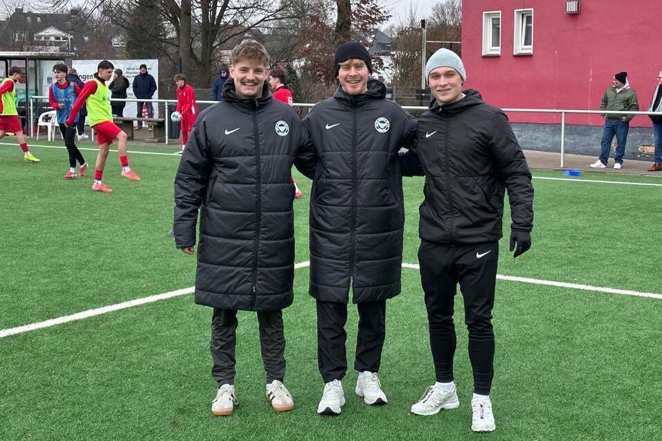 Drei junge M&auml;nner in Sportjacken stehen l&auml;chelnd auf einem Fu&szlig;ballplatz mit gr&uuml;nem Kunstrasen.