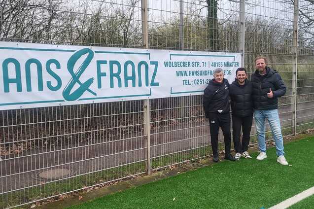 Drei Personen stehen vor einem Zaun mit einem Banner von "HANS & FRANZ" auf einem Fu&szlig;ballplatz.