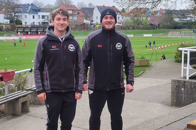 Zwei junge M&auml;nner in Sportjacken stehen auf einem Fu&szlig;ballplatz mit Spielern im Hintergrund.
