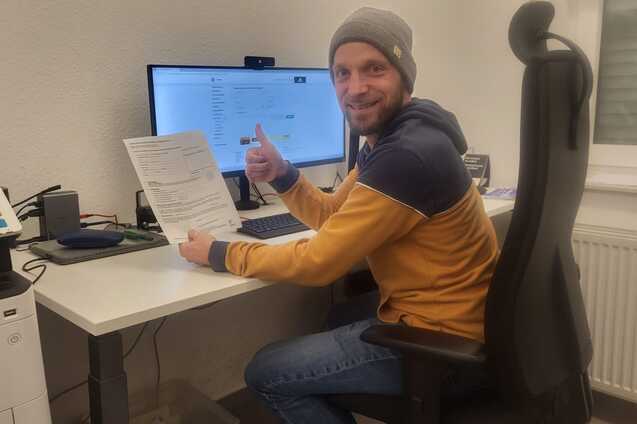Mann in einem B&uuml;ro zeigt Daumen hoch und h&auml;lt ein Dokument in der Hand, sitzt vor einem Computer mit Monitor.