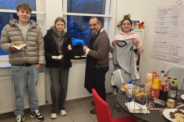 Vier Personen in einem Raum halten Essen und T-Shirts, w&auml;hrend einer eine Bestellung &uuml;bergibt. Hinter ihnen steht ein Tisch mit Snacks.