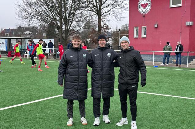 Drei junge M&auml;nner in Sportjacken stehen l&auml;chelnd auf einem Fu&szlig;ballplatz mit gr&uuml;nem Kunstrasen.