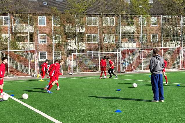 Gruppe von jungen Fu&szlig;ballspielern in roten Trikots trainiert auf einem Kunstrasenplatz mit zwei Toren im Hintergrund.