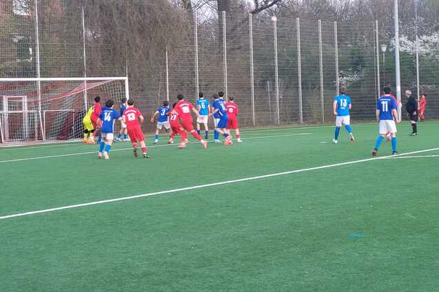 Spielszene auf einem Fu&szlig;ballplatz: Spieler in blauen und roten Trikots warten auf einen Freisto&szlig; in der N&auml;he des Tors.
