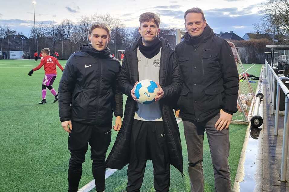 Drei Personen stehen auf einem Fu&szlig;ballplatz, eine h&auml;lt einen Ball, w&auml;hrend Spieler im Hintergrund aktiv sind.