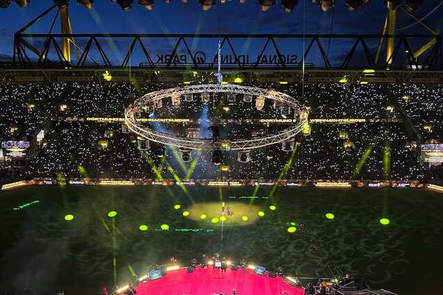 Luftaufnahme eines Stadions bei Nacht mit leuchtenden Lichtern und einer zentralen B&uuml;hnenkonstruktion auf dem Spielfeld.